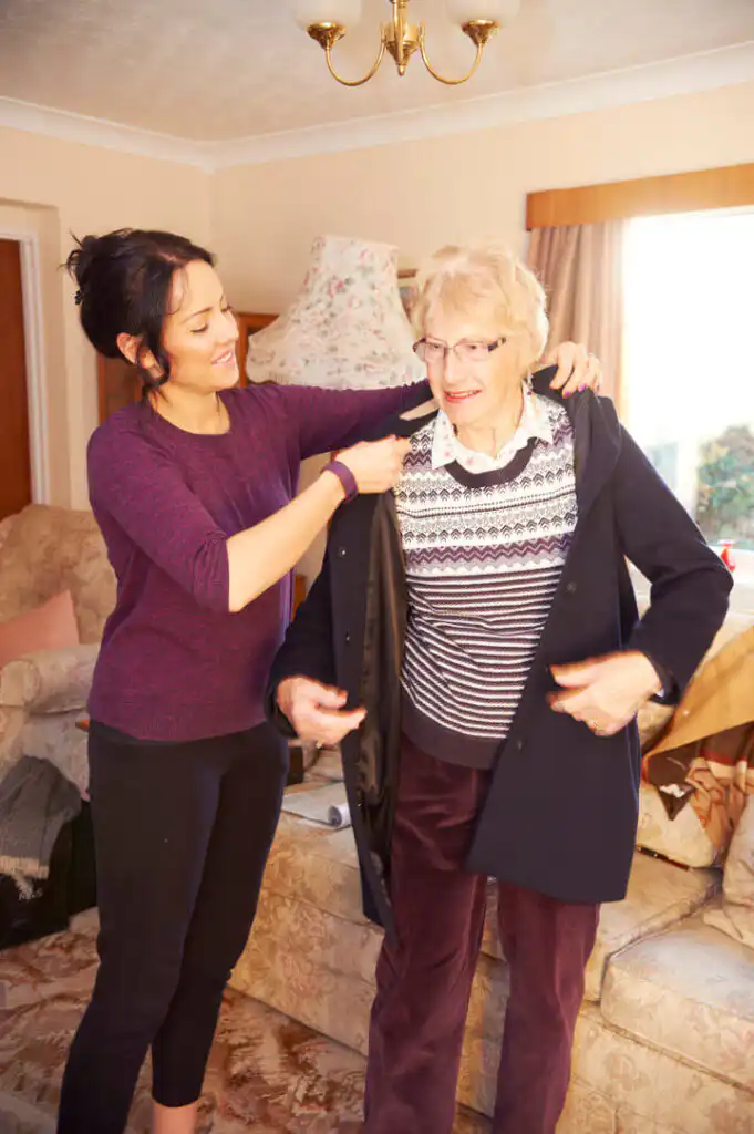 A young woman assists an older woman in putting on a coat in a cosy living room.