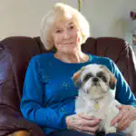 Older woman sat in a cosy brown armchair with a small white dog sat on her lap.