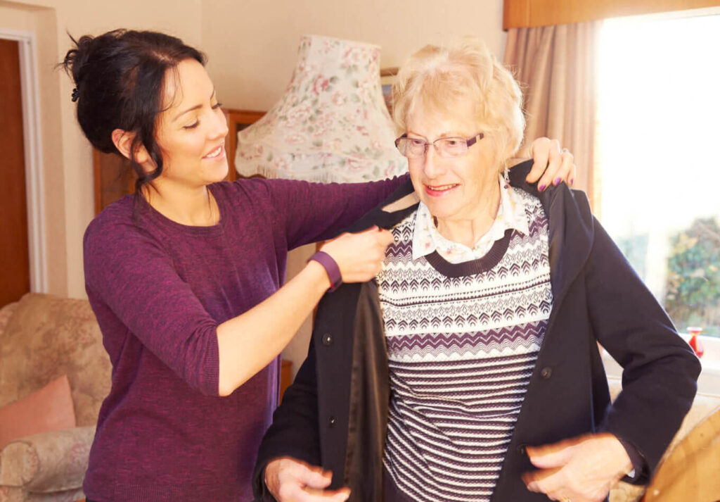 A young woman assists an older woman in putting on a coat in a cozy living room.
