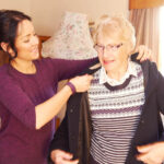 A young woman assists an older woman in putting on a coat in a cozy living room.