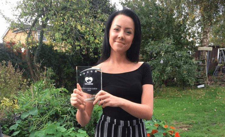 A woman with dark hair stands in a garden, smiling and holding a clear award plaque. She wears a black top and striped pants.