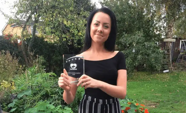 A woman with dark hair stands in a garden, smiling and holding a clear award plaque. She wears a black top and striped pants.