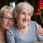 A smiling elderly woman and a woman with glasses share a warm embrace, sitting beside red tulips in a vase, conveying love and togetherness.