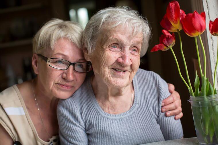 A smiling elderly woman and a woman with glasses share a warm embrace, sitting beside red tulips in a vase, conveying love and togetherness.