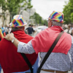 Two older adults wearing rainbow hats and red sweaters, with one arm around the other, walk down a street during a pride event.