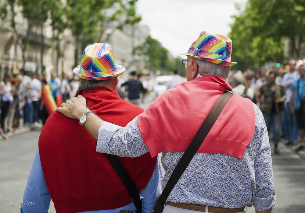 Two older adults wearing rainbow hats and red sweaters, with one arm around the other, walk down a street during a pride event.