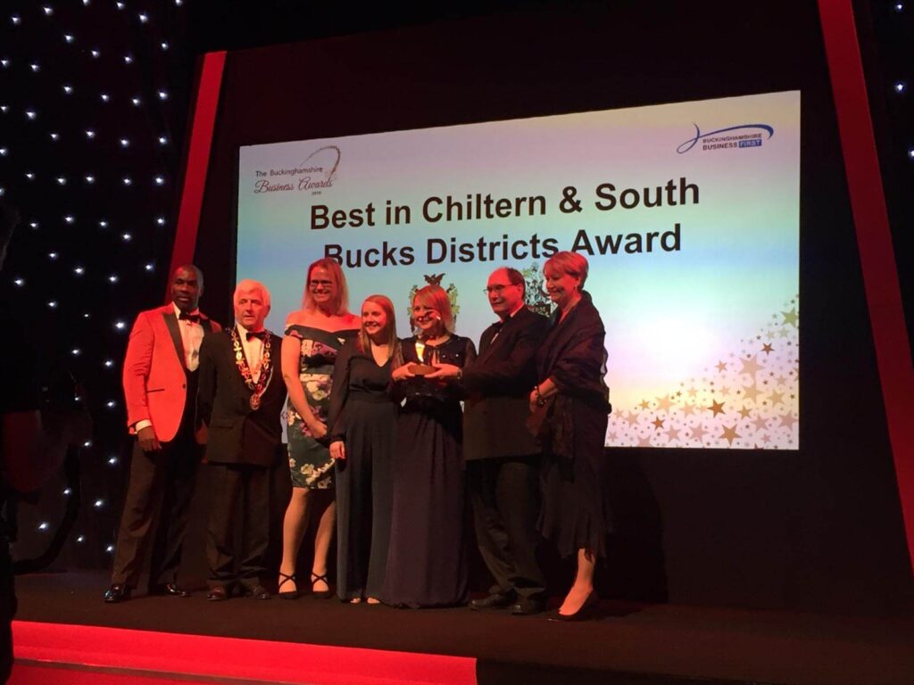 A group on stage smiles while holding an award. The backdrop reads "Best in Chiltern & South Bucks Districts Award" at the Buckinghamshire Business Awards.