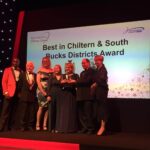 A group on stage smiles while holding an award. The backdrop reads "Best in Chiltern & South Bucks Districts Award" at the Buckinghamshire Business Awards.