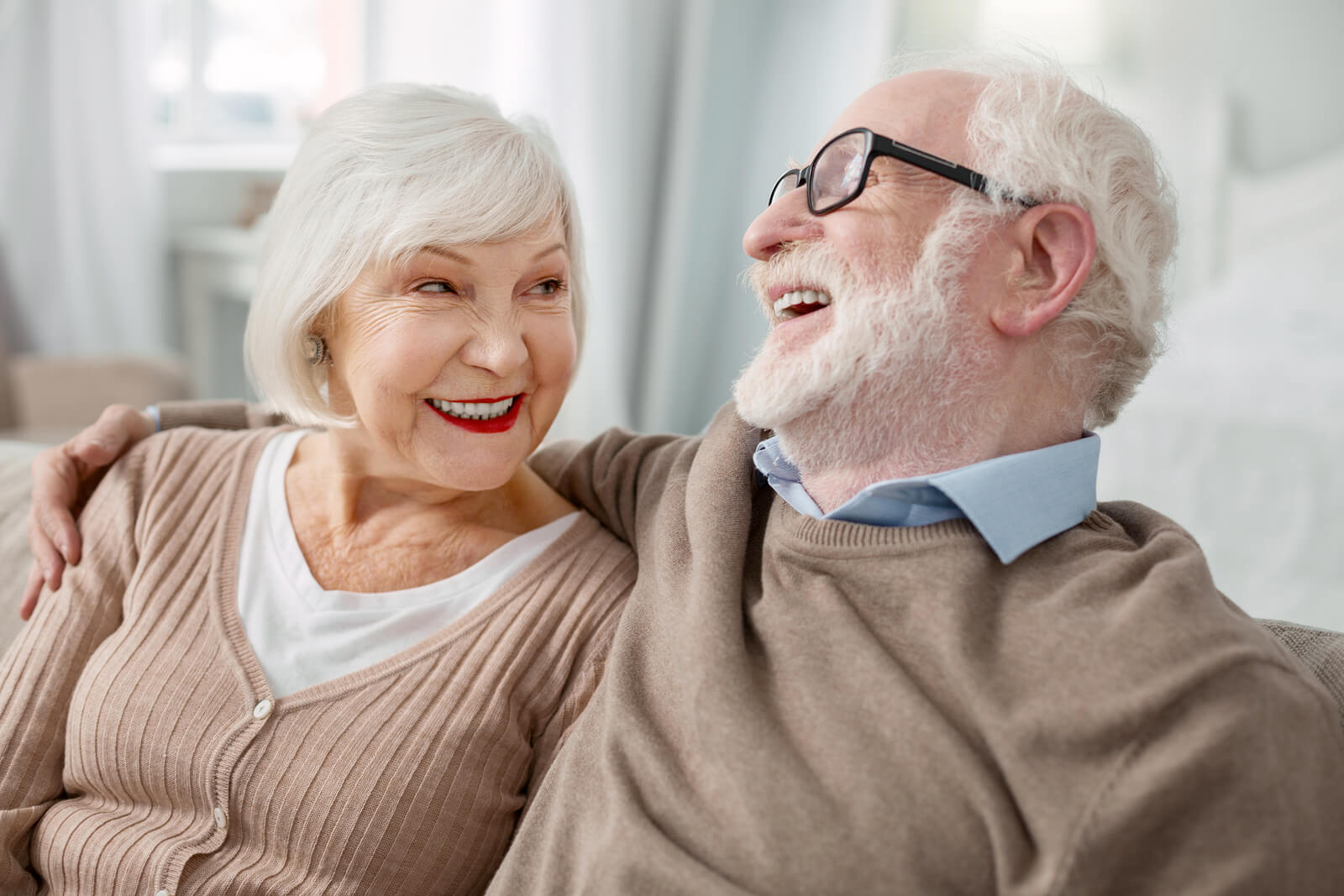 Elderly couple sitting closely on a sofa, laughing and gazing at each other warmly. Both wear glasses and cozy sweaters, conveying joy and companionship.