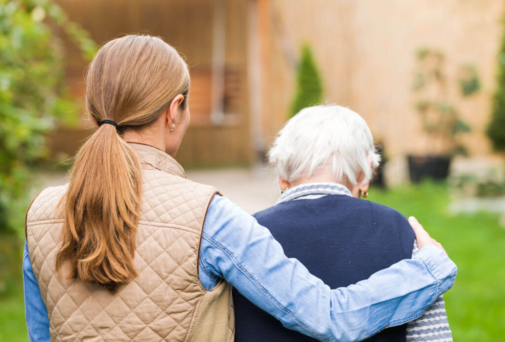 A woman with a ponytail, wearing a quilted vest, supports an elderly person in a garden.