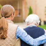 A woman with a ponytail, wearing a quilted vest, supports an elderly person in a garden.
