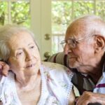 Elderly couple sitting close in a warmly lit room, the man gently hugging the woman as they both smile. Sunlight filters through a window behind them.