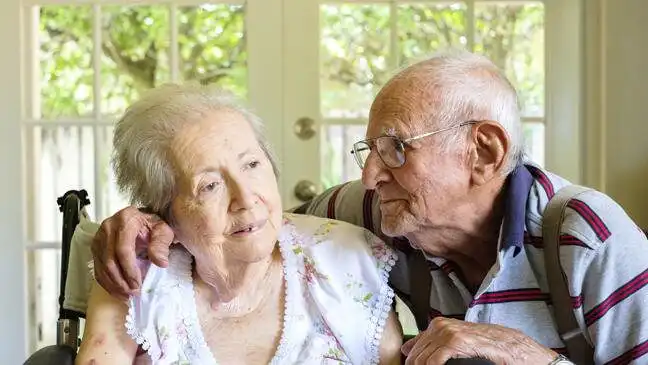 Elderly couple sitting close in a warmly lit room, the man gently hugging the woman as they both smile. Sunlight filters through a window behind them.