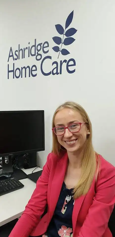 Client services manager Agata sat at her desk smiling at the camera. Tee Ashridge home care logo is visible on the white wall behind her.