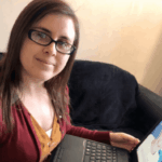 A woman with glasses and straight brown hair is smiling at the camera while sitting on a sofa, using a laptop. She wears a maroon cardigan and yellow top.