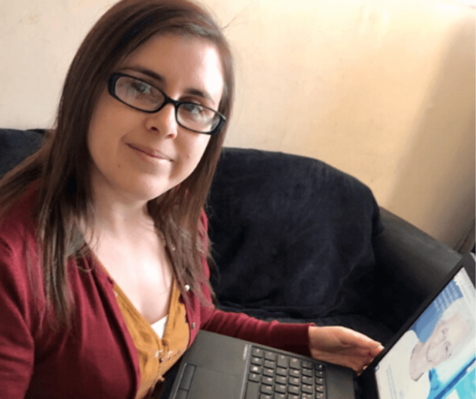 A woman with glasses and straight brown hair is smiling at the camera while sitting on a sofa, using a laptop. She wears a maroon cardigan and yellow top.