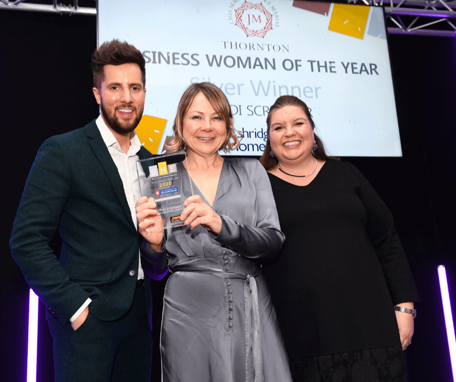 Three people smiling at an award ceremony. A woman in a grey dress holds a trophy, with a man and another woman standing beside her against a lit backdrop.