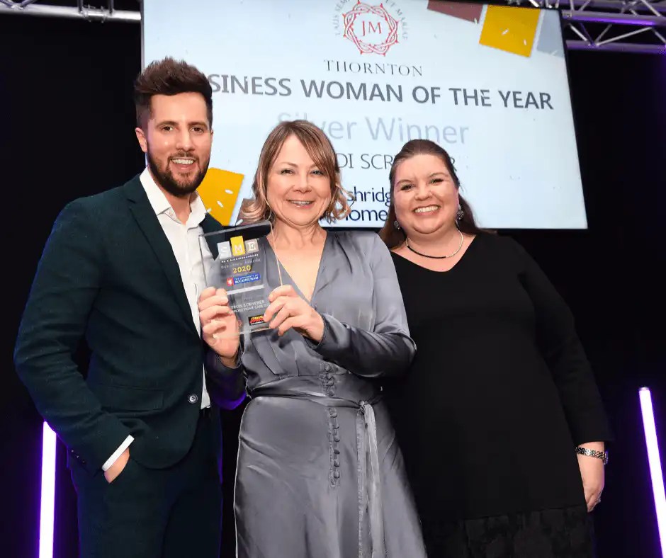 Three people smiling at an award ceremony. A woman in a grey dress holds a trophy, with a man and another woman standing beside her against a lit backdrop.