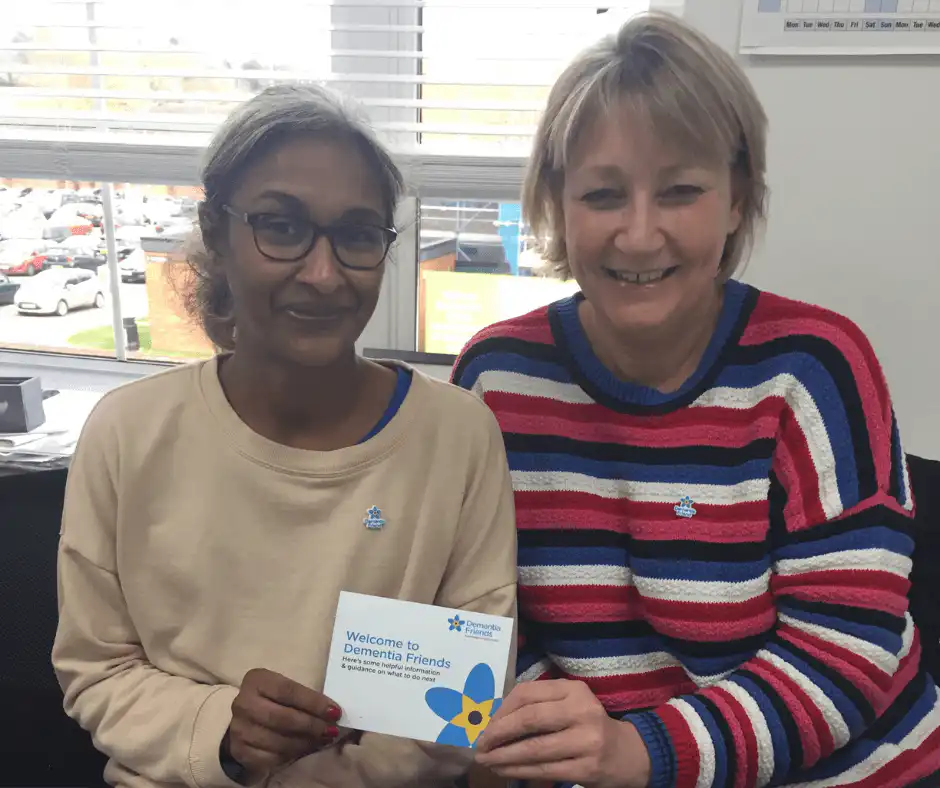 Two professional carers sat side by side smiling at the camera with a certificate that mentions they are now dementia friends.