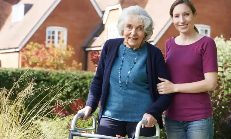 Older woman with walking frame standing in a garden in front of her house. A younger woman is stood next to her with a hand on her arm in a kind and supportive gesture.