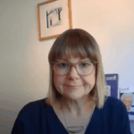 A person with glasses and a blue top smiles slightly in front of a white wall. Framed art pieces are hanging and standing on a shelf behind them.