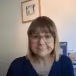 A person with glasses and a blue top smiles slightly in front of a white wall. Framed art pieces are hanging and standing on a shelf behind them.