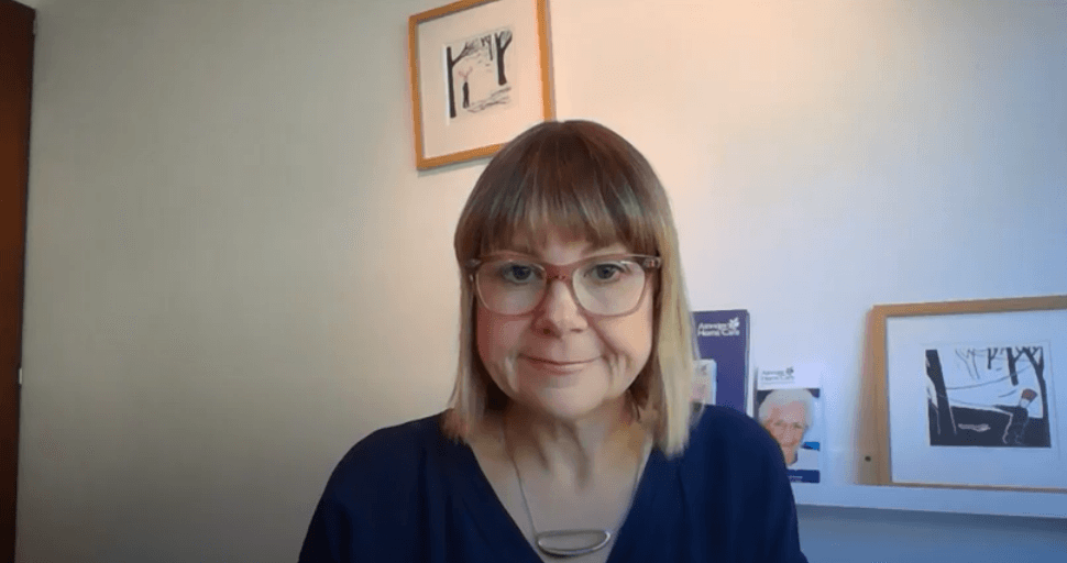 A person with glasses and a blue top smiles slightly in front of a white wall. Framed art pieces are hanging and standing on a shelf behind them.