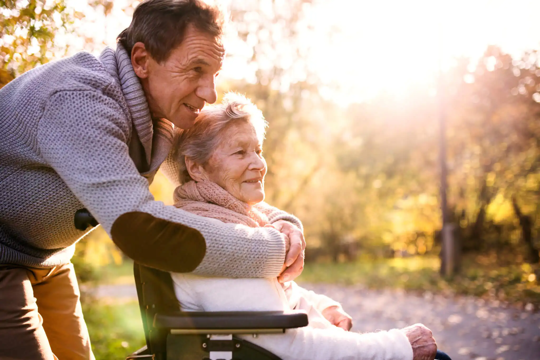 Older lady in a wheelchair being embraced by her husband, they are both smiling and staring off into the distance.