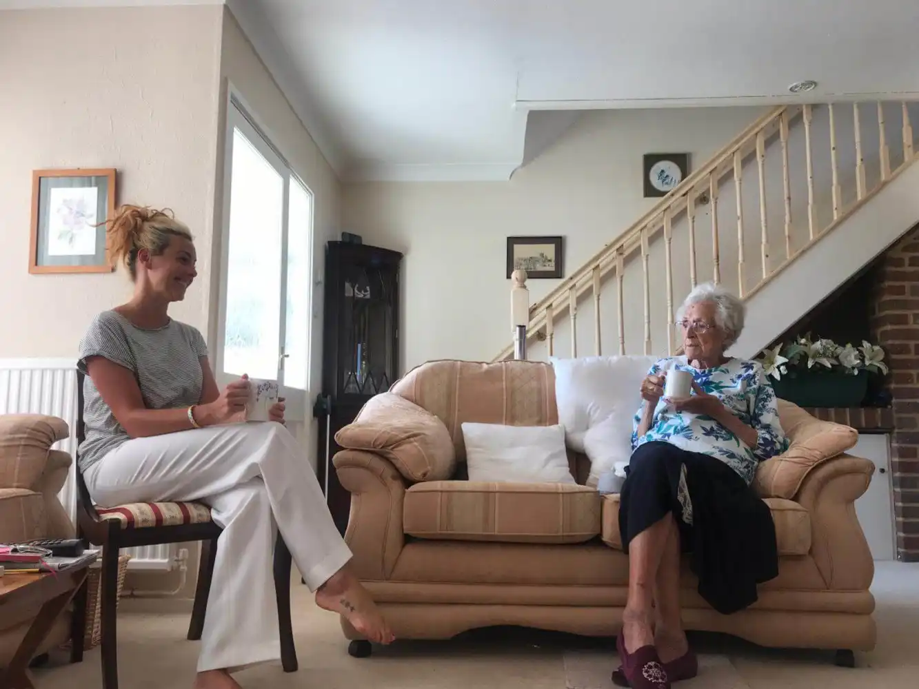 A young woman and an elderly woman sit in a cosy living room, holding mugs. They smile at each other, conveying warmth and connection.