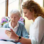 Older woman sitting next to a younger woman and being shown something on a handheld tablet.