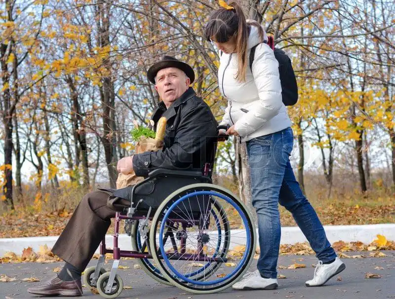 Older gentleman in a wheelchair being supported to go out and do his shopping by his carer.