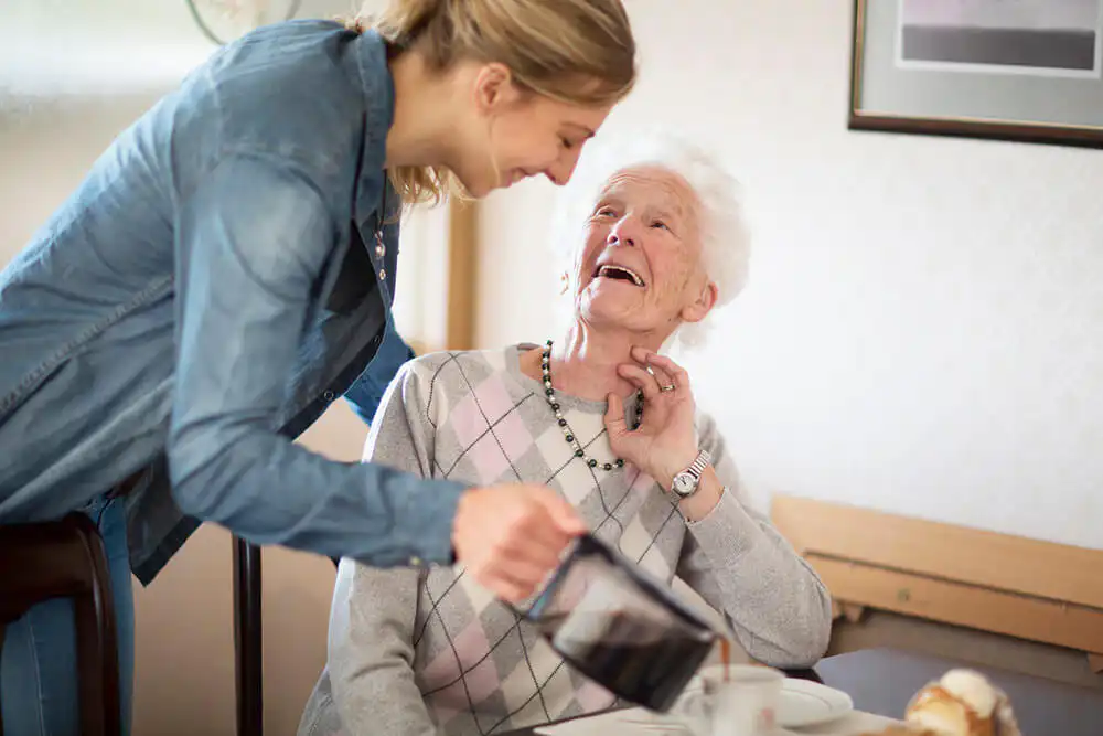 Older woman sitting at her dining table, smiling as her carer pours her some coffee.