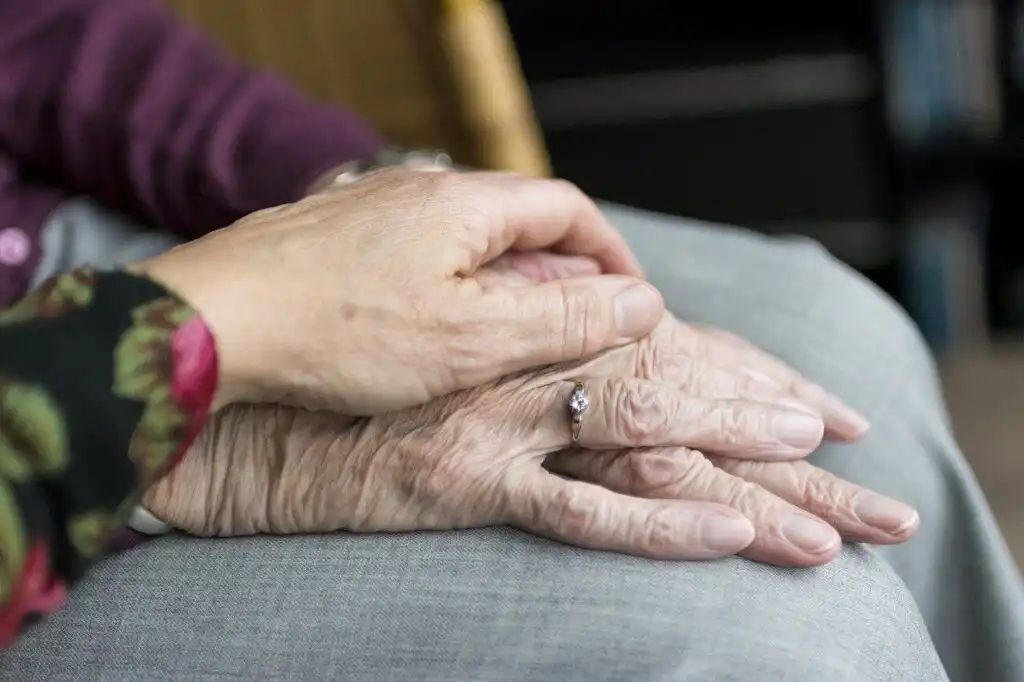 Close up of an elderly person resting their hands on their knee, with another person placing their hand on top in a comforting gesture.