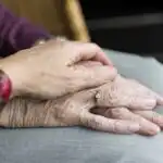 Close up of an elderly person resting their hands on their knee, with another person placing their hand on top in a comforting gesture.