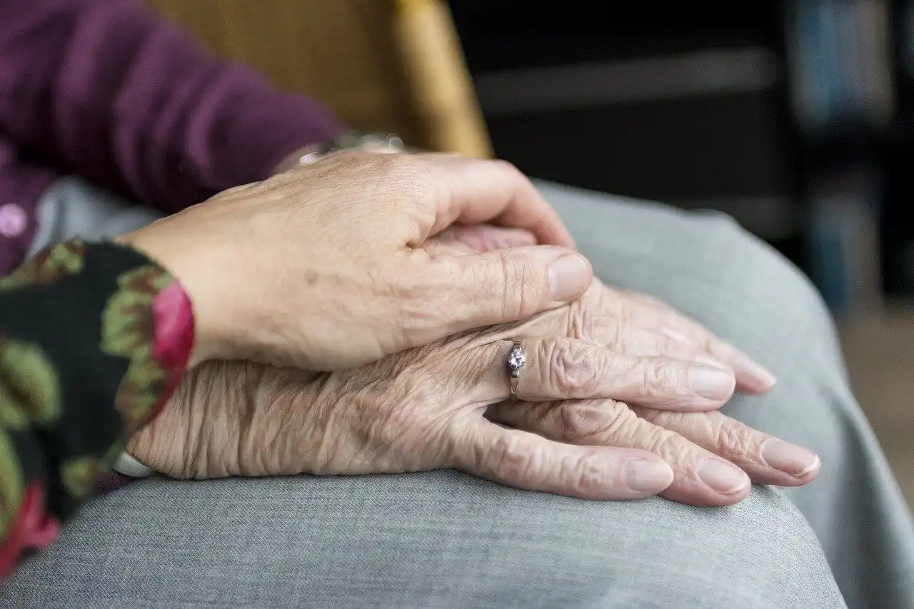 Close up of an elderly person resting their hands on their knee, with another person placing their hand on top in a comforting gesture.