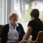 An elderly woman and a younger woman share a joyful conversation in a sunlit room. The older woman sits with a cane beside her, both smiling warmly.