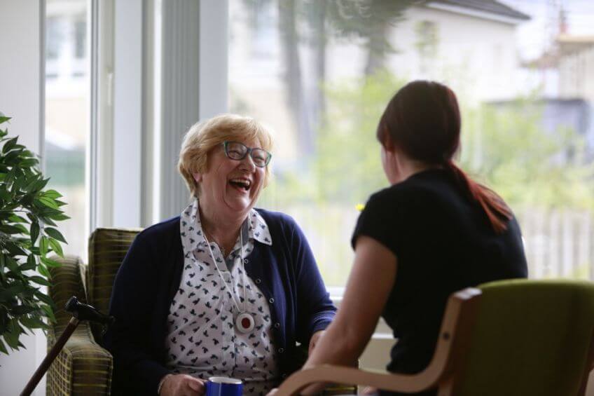 An elderly woman and a younger woman share a joyful conversation in a sunlit room. The older woman sits with a cane beside her, both smiling warmly.