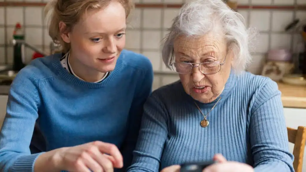Older lady sat at a kitchen table next to a younger lady, they are both looking at a mobile phone with interest.