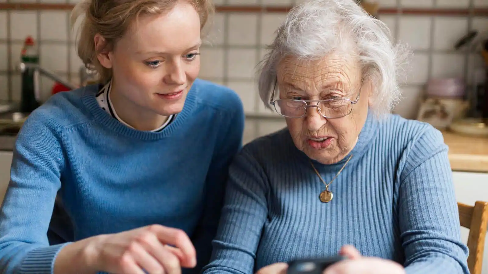 Older lady sat at a kitchen table next to a younger lady, they are both looking at a mobile phone with interest.