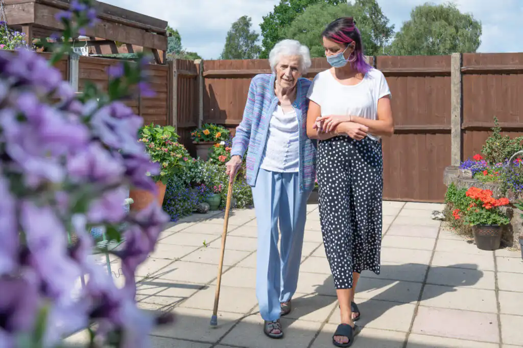 Elderly lady walking across the patio in her garden with a walking stick. She is supported by a younger carer who is holding her arm in a supportive, guiding manner. The carer is wearing a facemask and focusing on the elderly lady, whilst she look at the flowers in her garden.