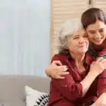 Older lady sitting on a sofa with a younger woman who has her arm around her. They are both smiling.