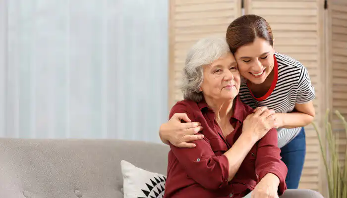 Older lady sitting on a sofa with a younger woman who has her arm around her. They are both smiling.