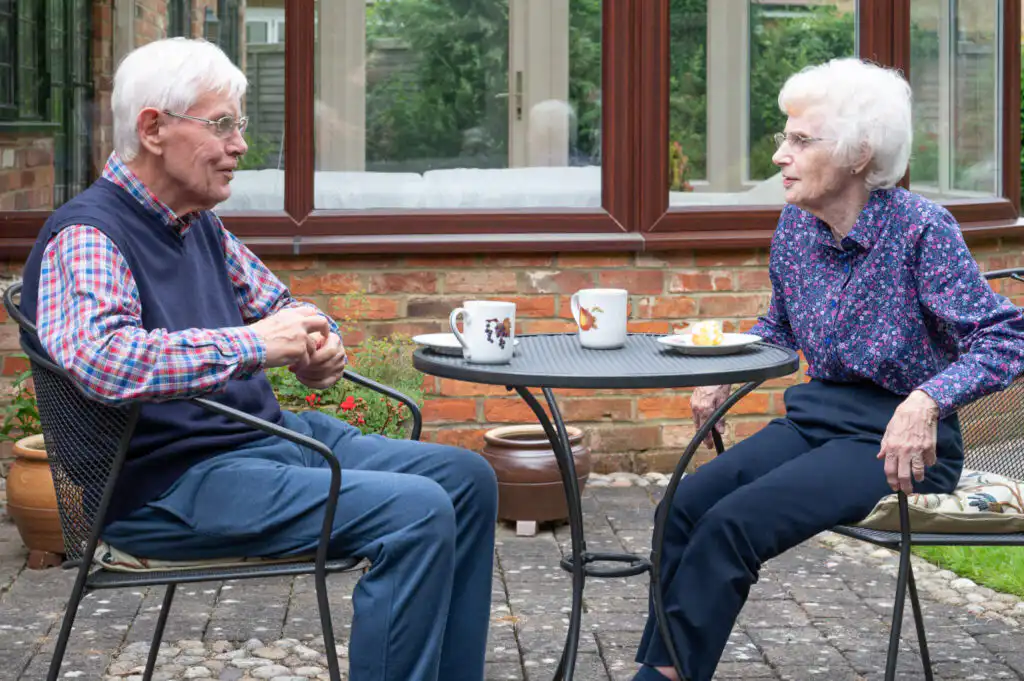 An elderly couple say enjoying tea and cake on the patio in their garden.