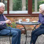 An elderly couple say enjoying tea and cake on the patio in their garden.