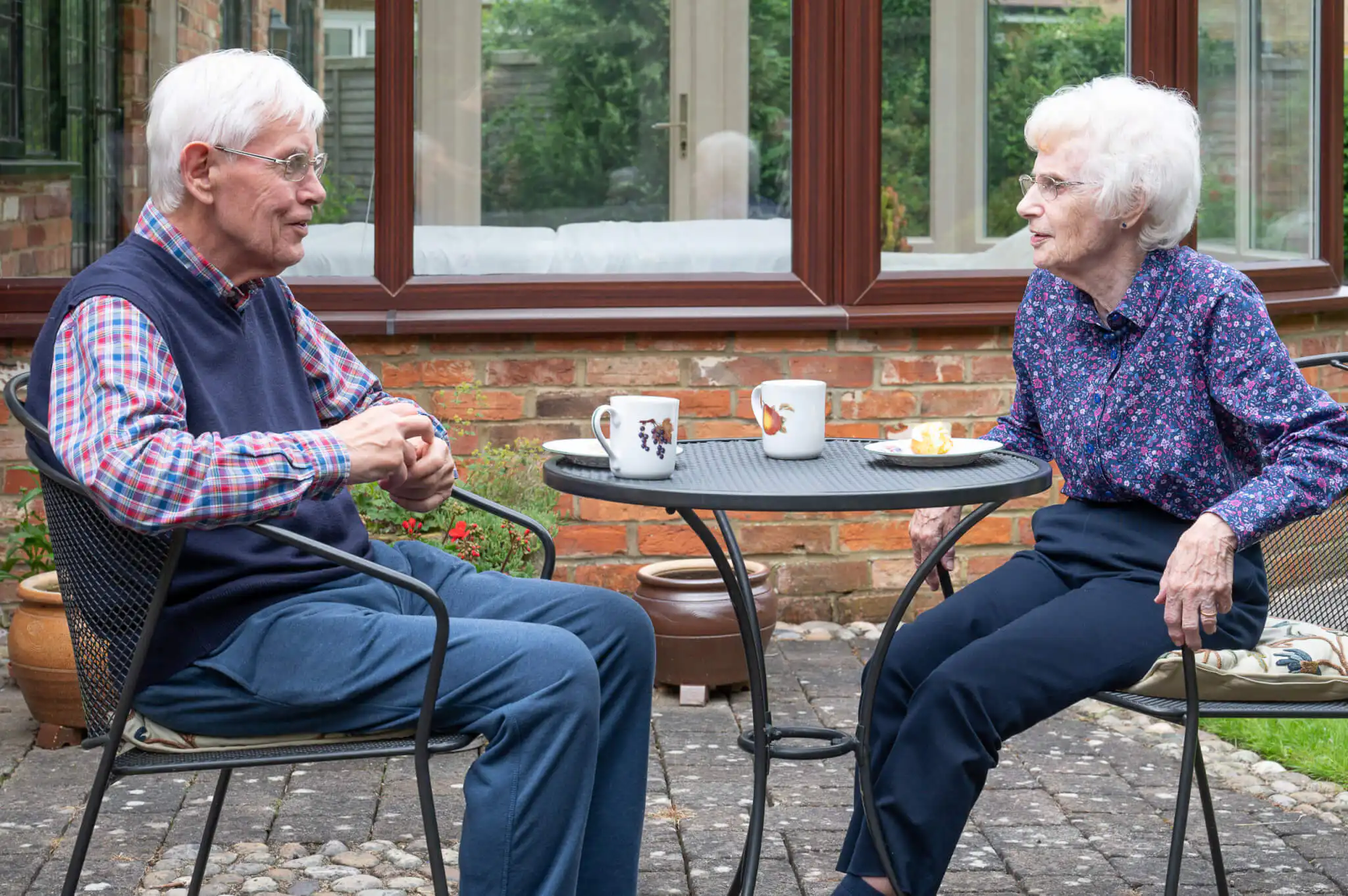 An elderly couple say enjoying tea and cake on the patio in their garden.