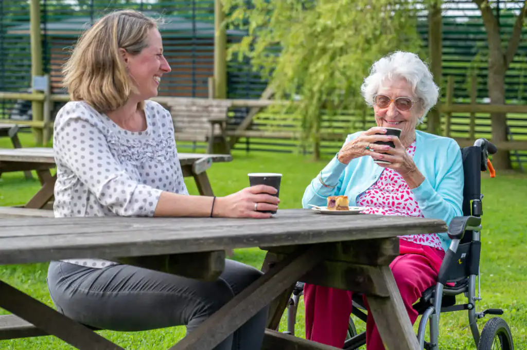 Elderly lady sitting with a professional carer at a picnic bench, enjoying a cup of tea and a slice of cake in the sunshine.