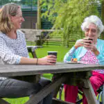 Elderly lady sitting with a professional carer at a picnic bench, enjoying a cup of tea and a slice of cake in the sunshine.
