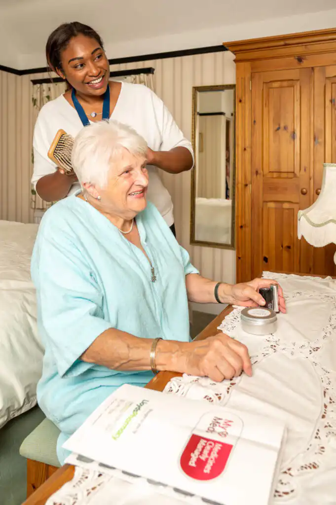 A smiling elderly woman in a light blue top sits at a table with papers, while a cheerful caregiver stands beside her, holding a book, in a cosy room.
