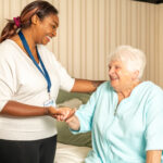 A caregiver and elderly woman share a joyful moment. The caregiver, smiling, holds the woman's hand as she sits on a bed in a cozy, well-lit room.