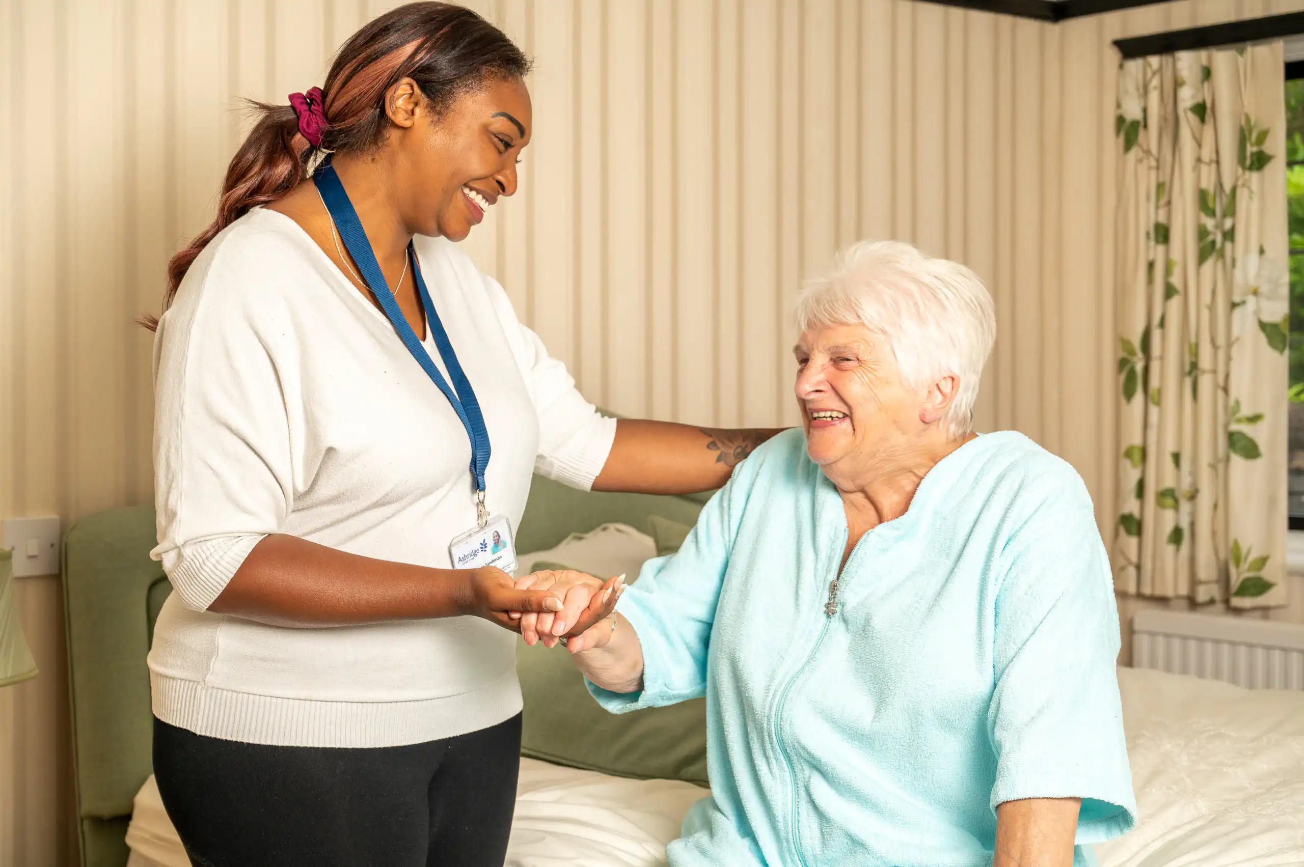 A caregiver and elderly woman share a joyful moment. The caregiver, smiling, holds the woman's hand as she sits on a bed in a cosy, well-lit room.
